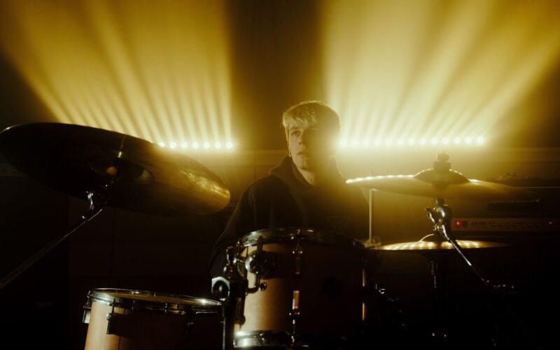 Baterista atrás da bateria, com fachos de luz amarela ao fundo